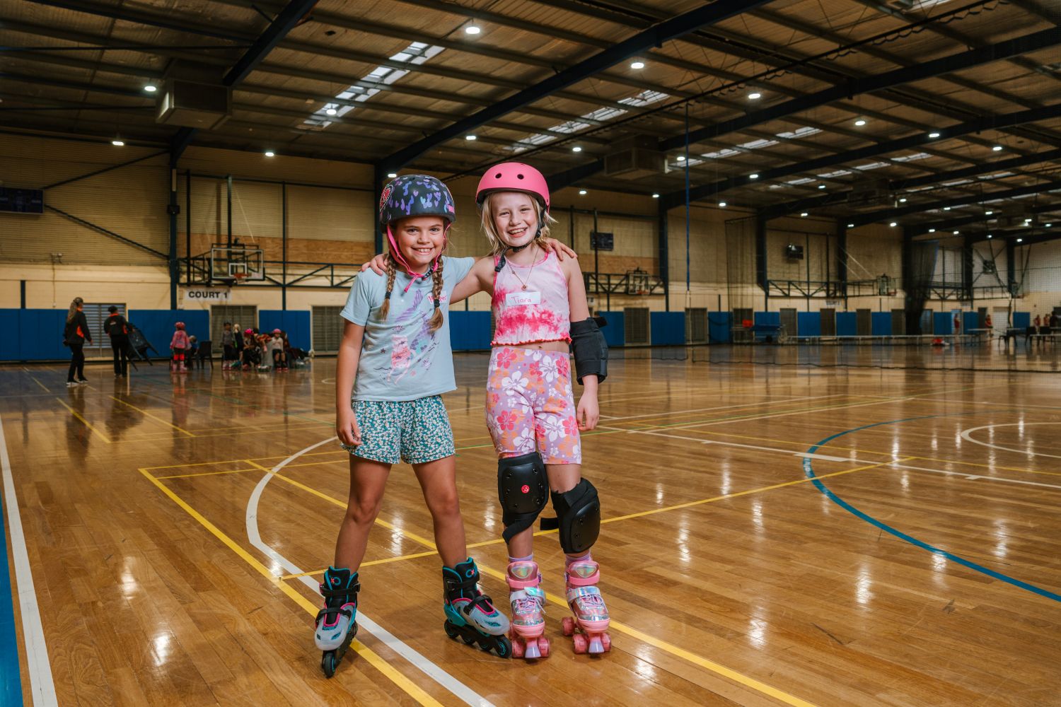 Two smiling young girls wearing helmets, knee pads, and roller skates stand arm-in-arm on a large indoor timber sports court with a high ceiling.
